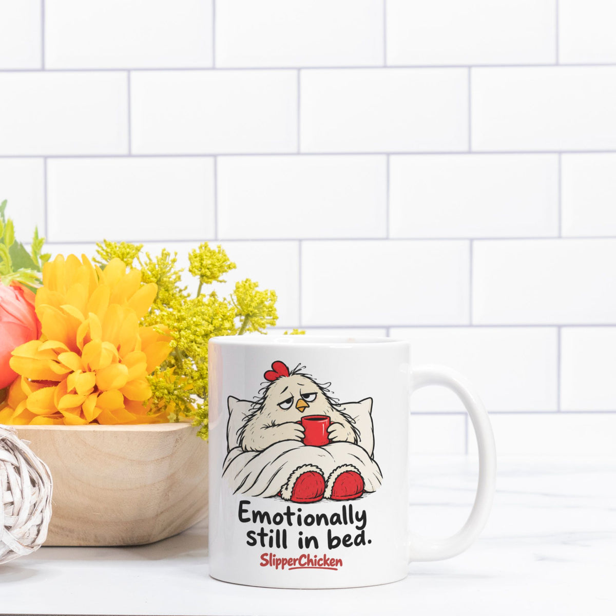 White Sleepy Mug with a chicken tucked into bed, Emotionally still in bed quote, and red SlipperChicken logo
